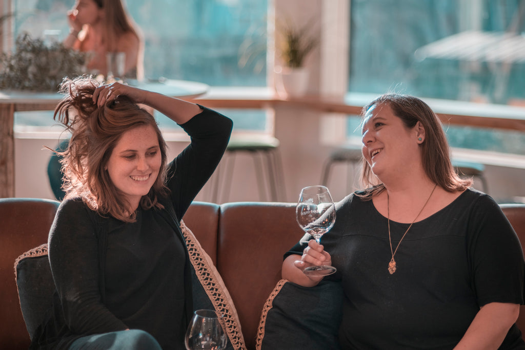 Allie and Julie sitting on a couch in a wine tasting room drinking wine and laughing